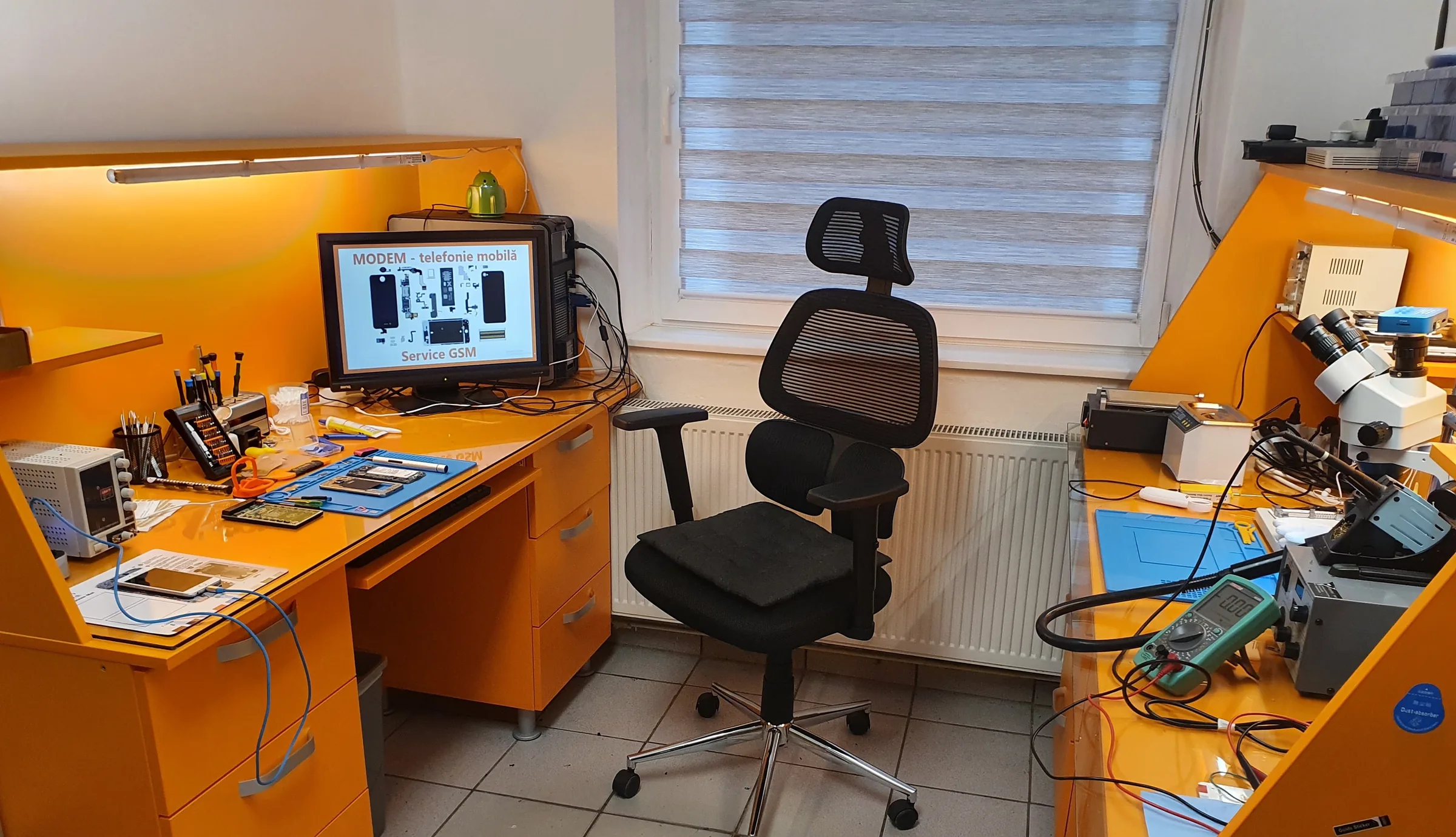 Atelierul de reparații MODEM: bancuri portocalii, microscop, stație de lipit, multimetru